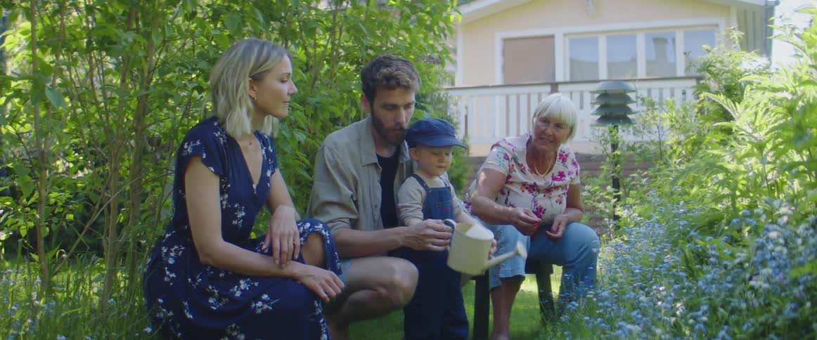 A family and grandmother kneel together outside while the small boy uses a watering can on some flowers