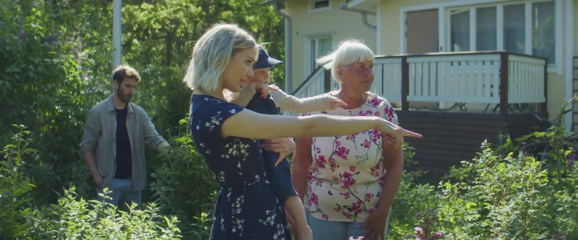 Mom holds her son and points at flowers and bushes in the garden while dad and grandma look on