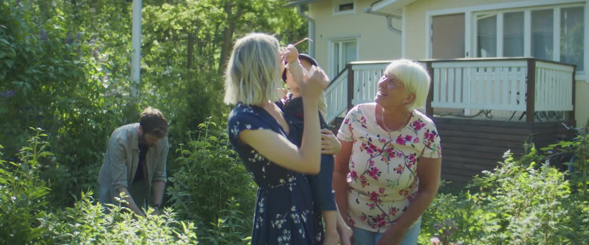 A mother father grandmother and grandson talk outside of a home while father does yard work