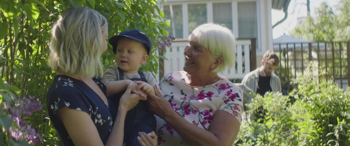 Mom and grandma laugh and kiss baby while standing in the garden on a sunny day