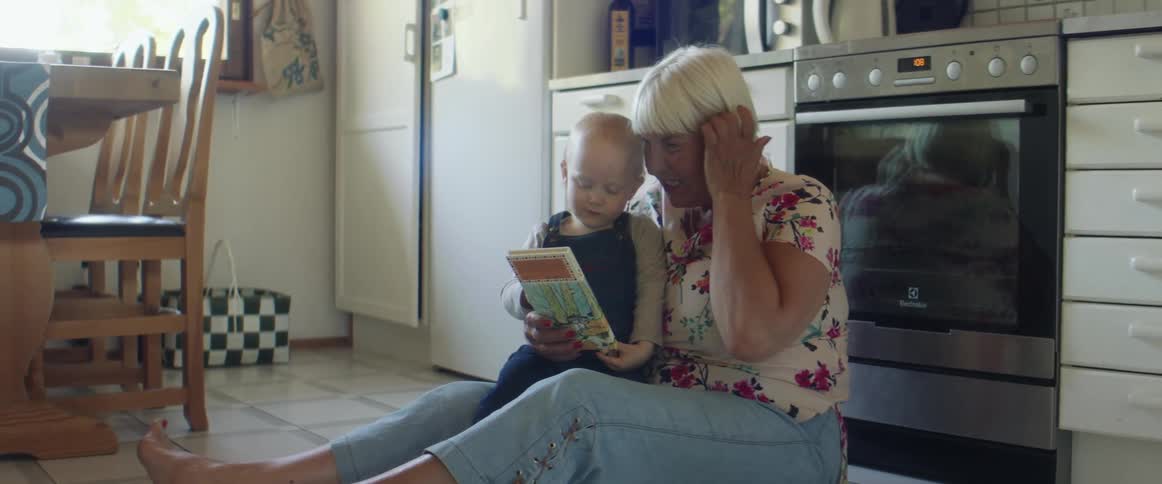 Grandma holds her arms out for a little boy and pulls him onto her lap as she reads a book while sitting on a kitchen floor