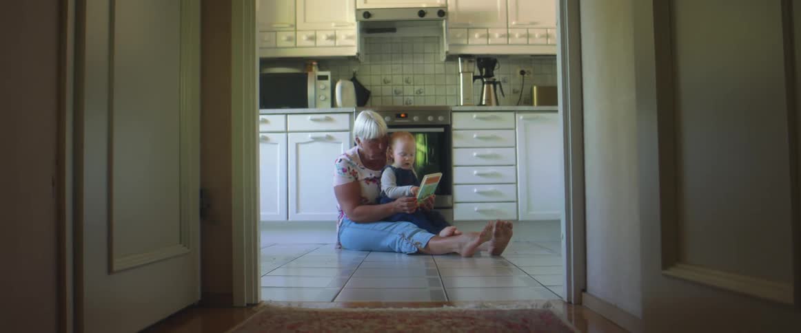 A woman sits on the floor of her kitchen and reads to her granddaughter