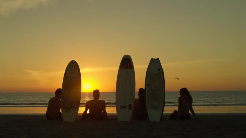 The silhouette of friends sitting in the sand with surfboards standing watching the waves at sunset