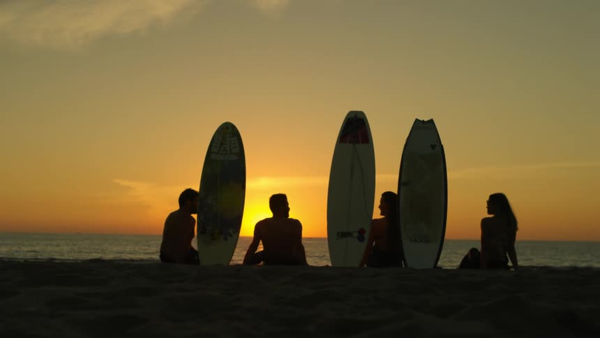 People sitting on beach near standing surfboards talk silhouetted by sun sitting on horizon in red sky