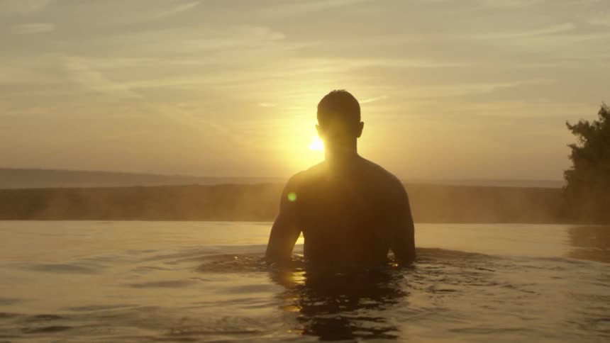 A silhouette of a man standing in water with the sunset the sky and clouds in the background