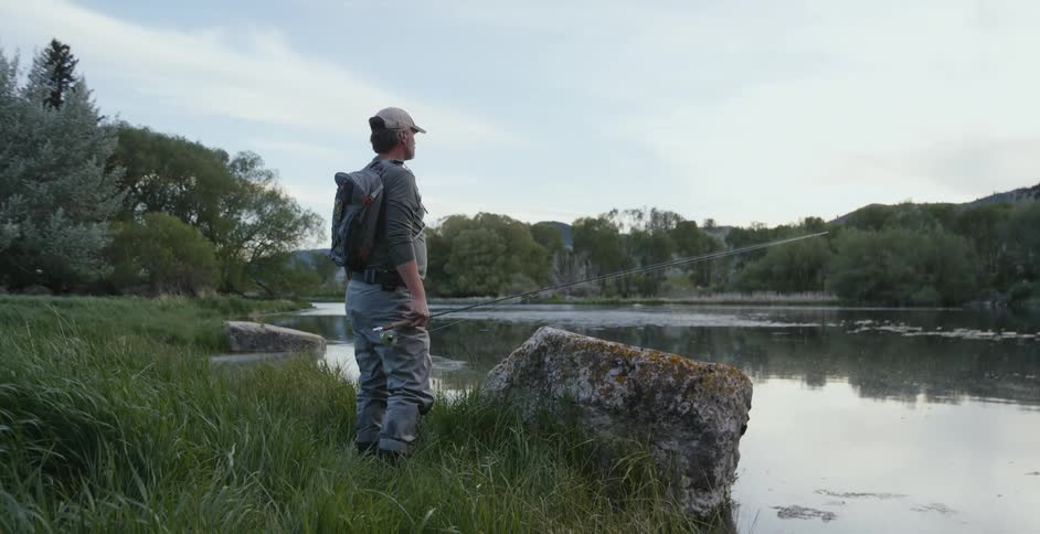 A man with a fishing pole looks out over a lake while standing on its shore during the day