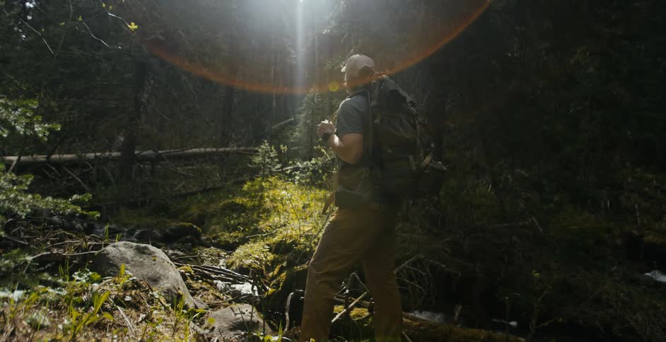 A man with a hat and a backpack standing in the forest next to a stream