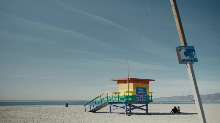 Two people sit in the shade of the lifeguard post on an empty beach on a bright sunny day
