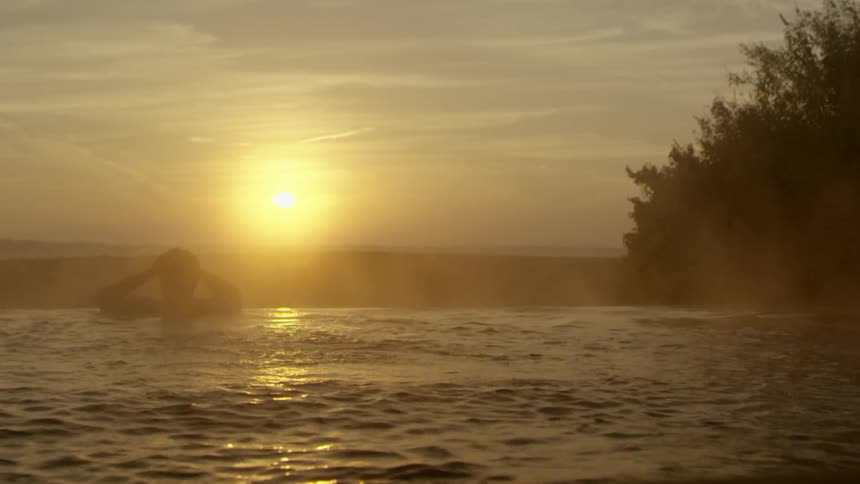 A man is swimming in the lake while the sun is setting