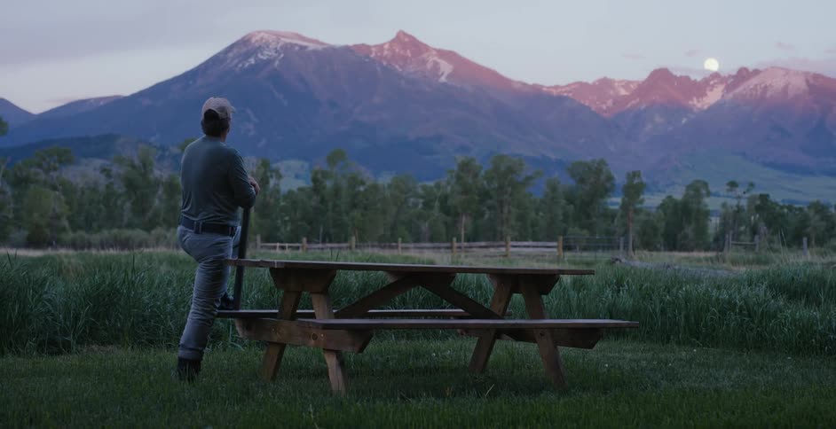 A man standing with a foot on the bench of a picnic table looks out at the distant mountains