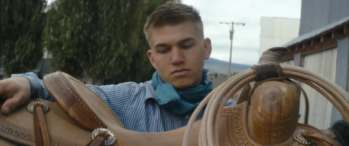 Man sets saddle over saddle blanket with ranch house and horizon in background