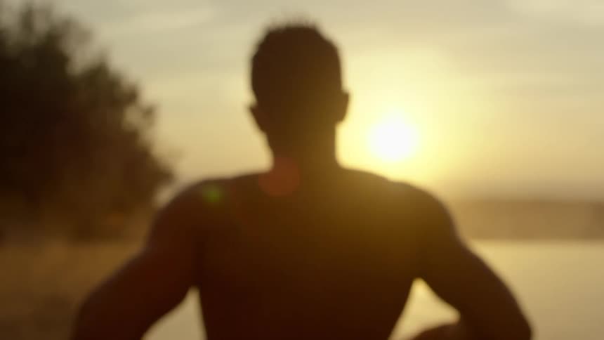 Silhouette of a strong muscular shirtless man standing in front of the ocean at sunset