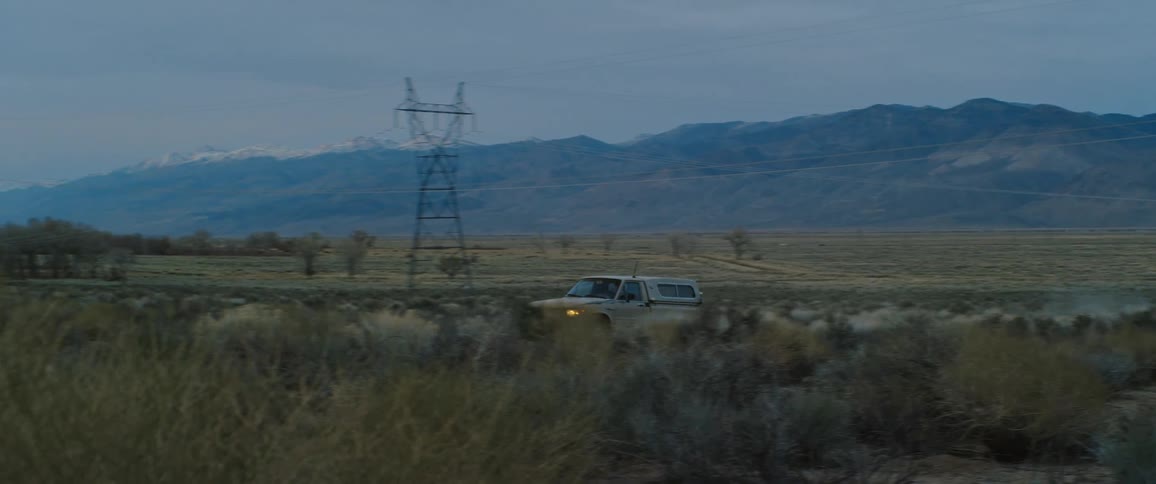A truck with headlights on drives on a road lined with utility towers and mountains in the distance