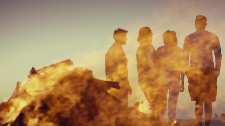 Four people stand on a beach in front of a bonfire