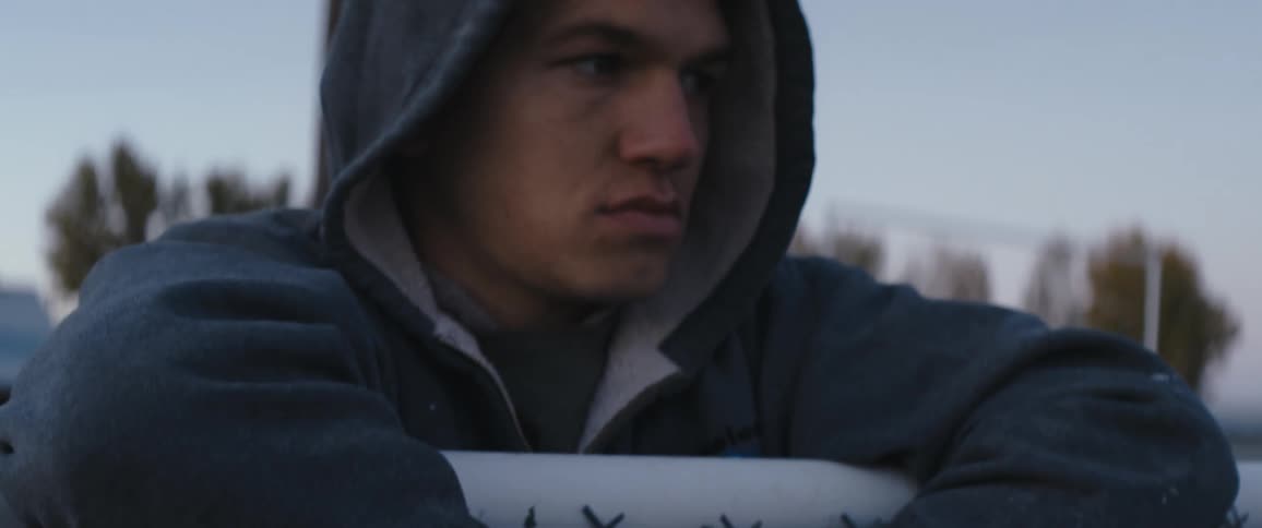 Young man wearing a hoodie reclined over a wire fence outdoors at evening