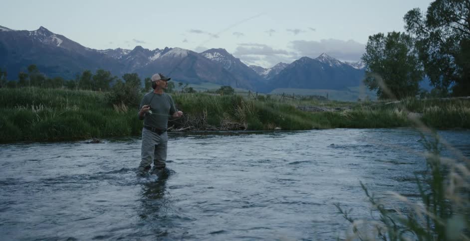 A man stands in a river and throws his fishing line out to the water