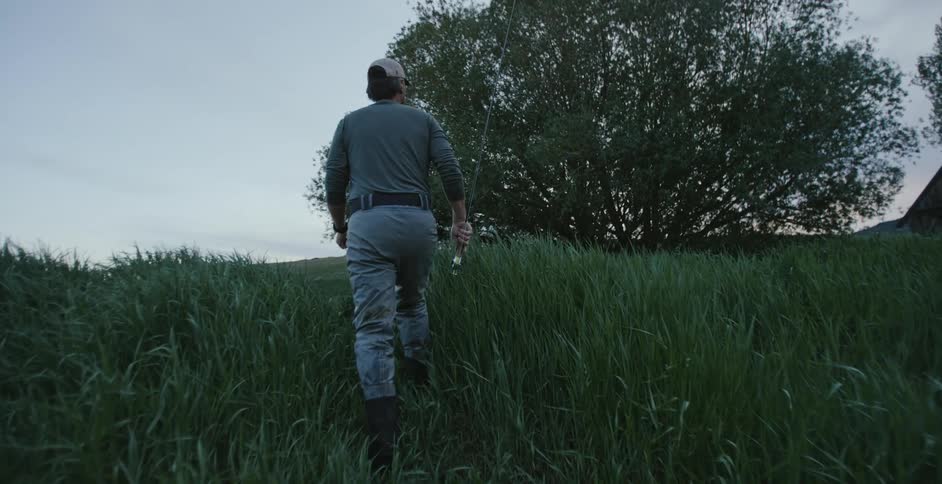 A man is walking through tall grass carrying a fishing rod and headed toward a camper