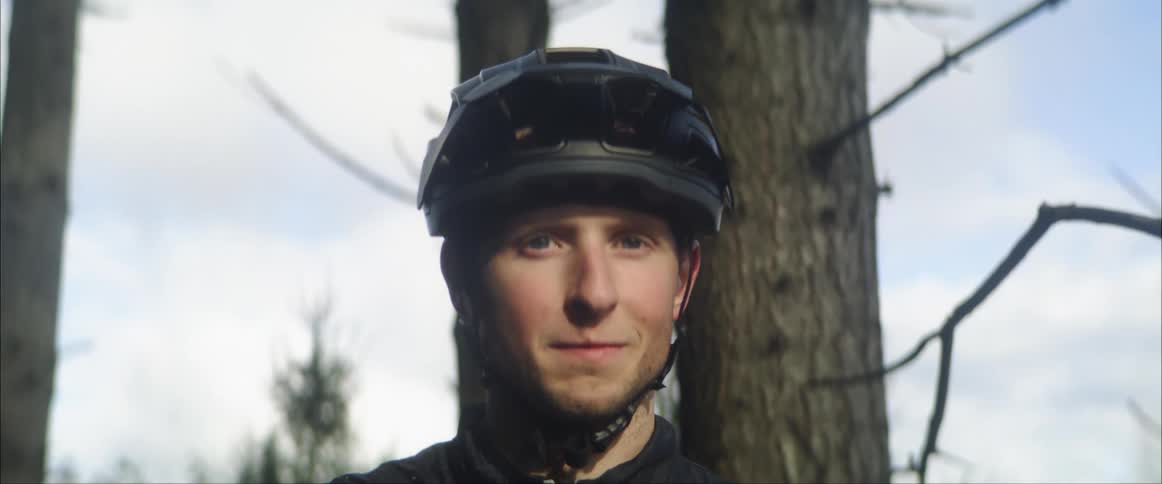 A male bicyclist who is standing outdoors slowly looks upwards as he poses for the camera