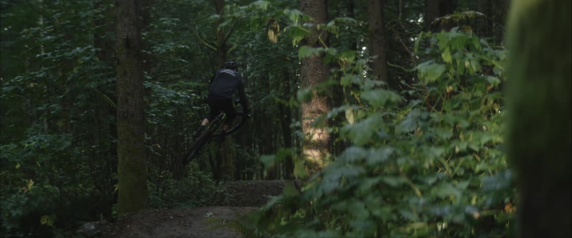 A cyclist goes airborne after riding up a dirt ramp on a dirt trail in a woodland and lands further down the trail