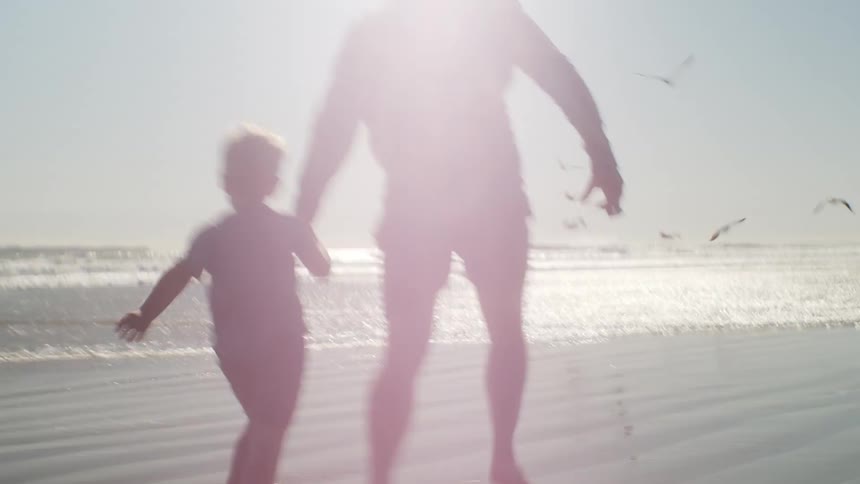 Father and son run hand in hand along wet sand into surf as seabirds take to the air and fly