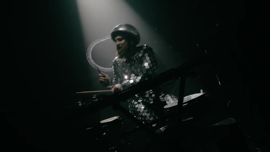 A male drummer vigorously strikes his drums under flashing strobe lights and in front of a screen with abstract imagery projected onto it