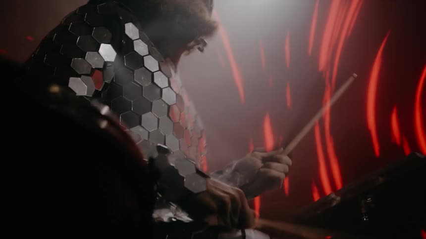 A drummer performs in a silver sequined costume as swirled reds lines glow behind him