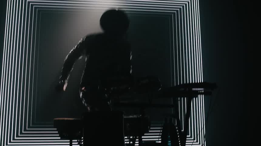 A drummer in a sequin suit stands up after playing in front of a patterned screen