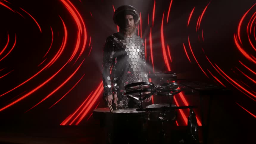 A male drummer wearing a strange reflective outfit stands in front of a projection of lights and standing at a drumkit