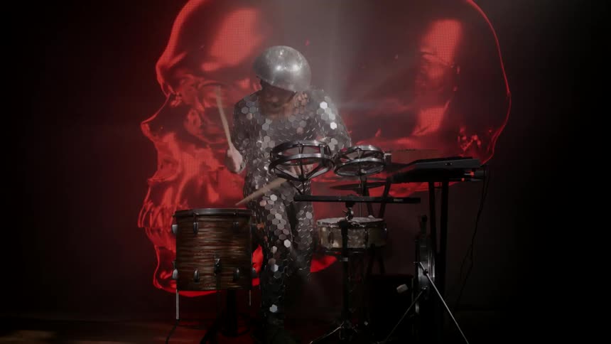 A drummer in a glittering bodysuit and helmet plays the drums in front of a giant rotating metallic skull