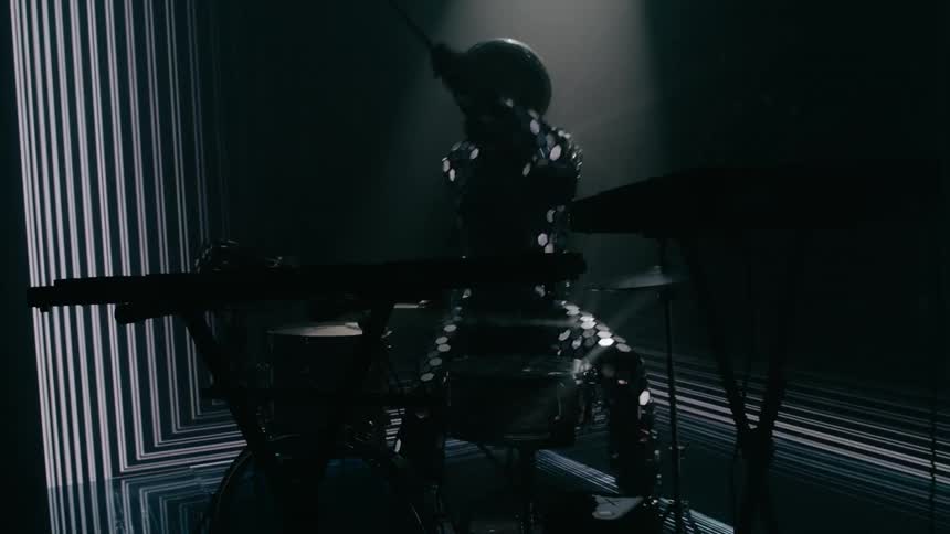 A drummer wearing a sequin suit and helmet plays while a light show strobes and flashes behind and around him