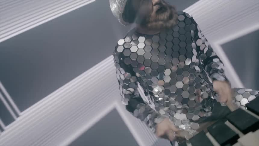A man with a beard is dressed in a mirror suit and playing the drums while lights strobe and flash