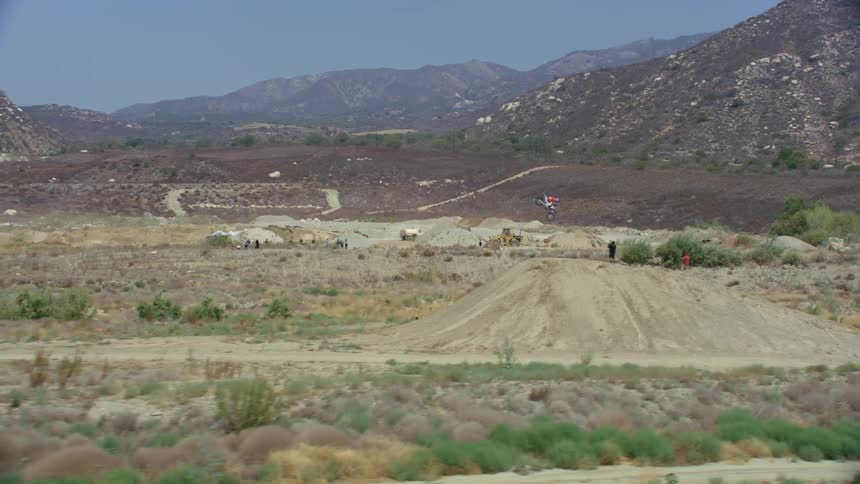 A person on an offroad motorcycle rides up a long ramp and flips in the air before landing on a large dirt mound