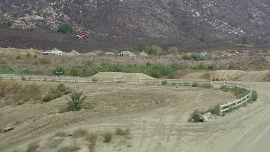 A aerial view of a dirt bike competition where a biker makes a high jump over a road with mountains in the background