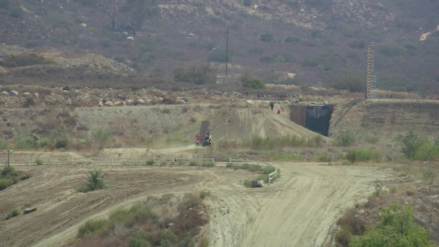 Person rides up ramp set to leap toward mound other people wait on with mountain slope and scrub in background