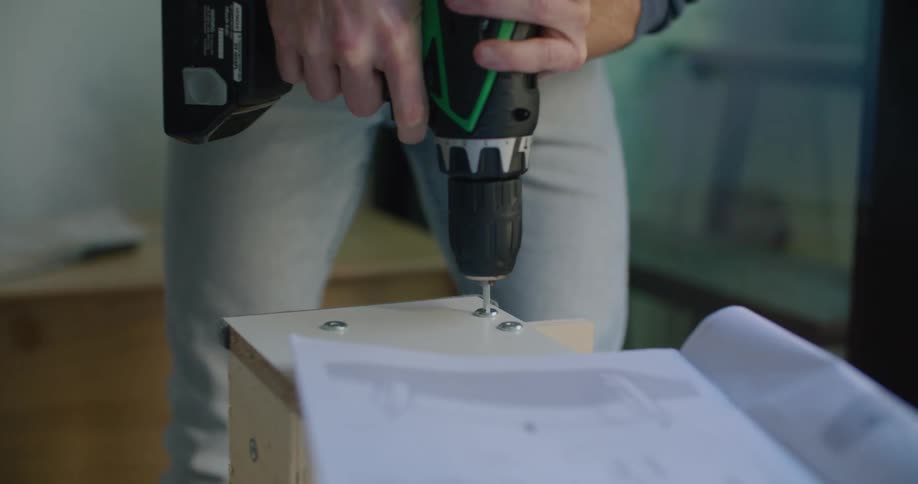 A male stands over a piece of furniture that he is assembling using a power tool