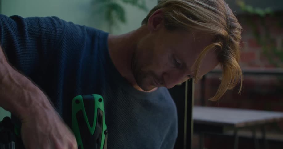 A man using a power drill on a piece of furniture with a table and a wall in the background