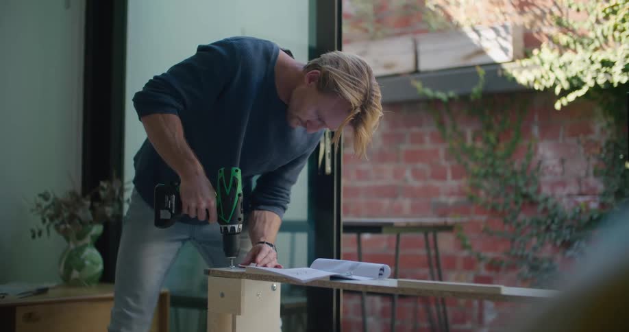 A man uses a power drill to put in a screw while reading assembly instructions
