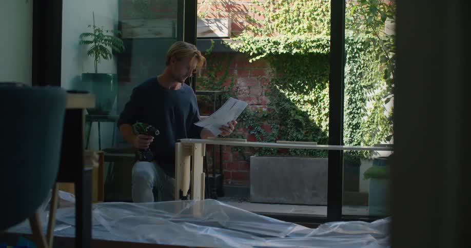 A man stops to read a manual while using a cordless drill to build furniture in his living room