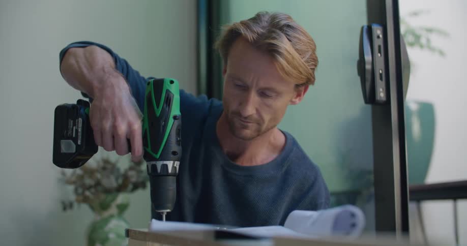 A man checks an instruction manual while assembling furniture with a power drill