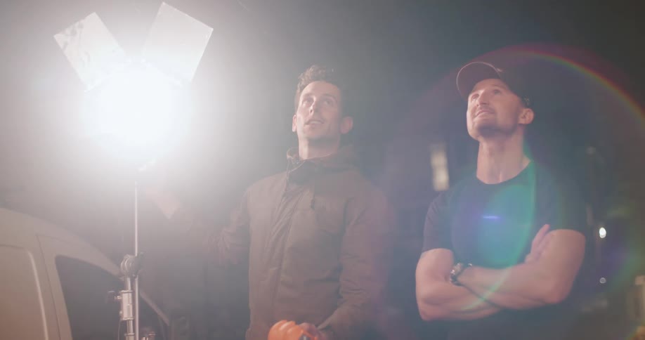 Two men stand outside at night as one turns on a bright light in preparation for a photo shoot