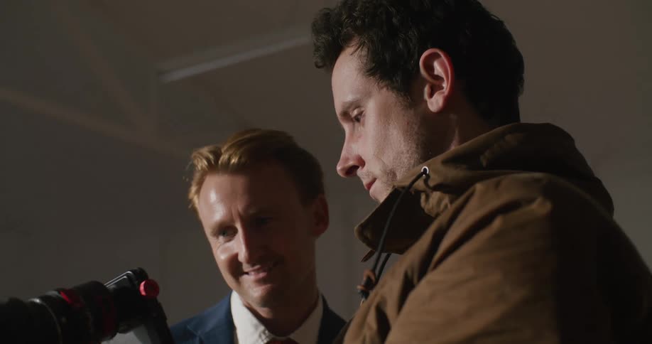 Two men in a room looking at a camera with a person and a wall in the background