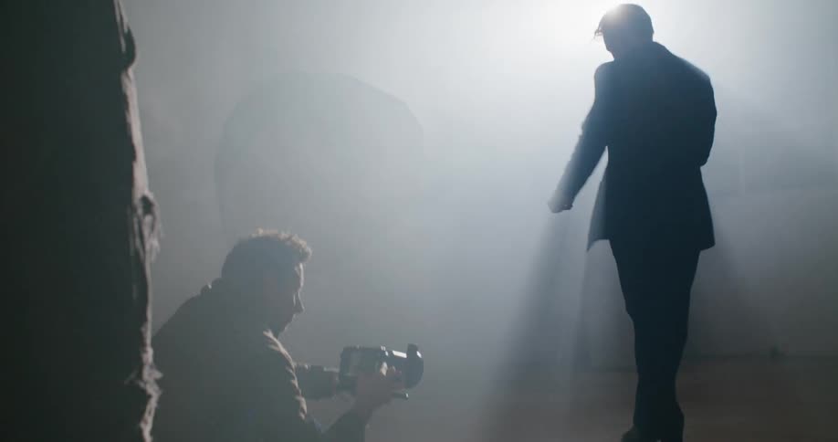 A man sitting on the floor with a video camera filming a man dancing in a room