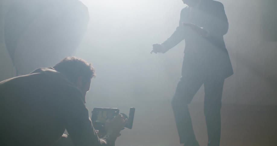 A man in a room with a camera filming a man dancing with a wall in the background