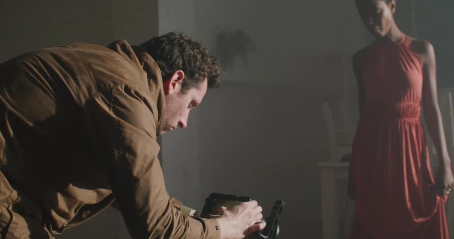 A man with a camera in a room filming a woman with a dress with a wall tables and chairs in the background