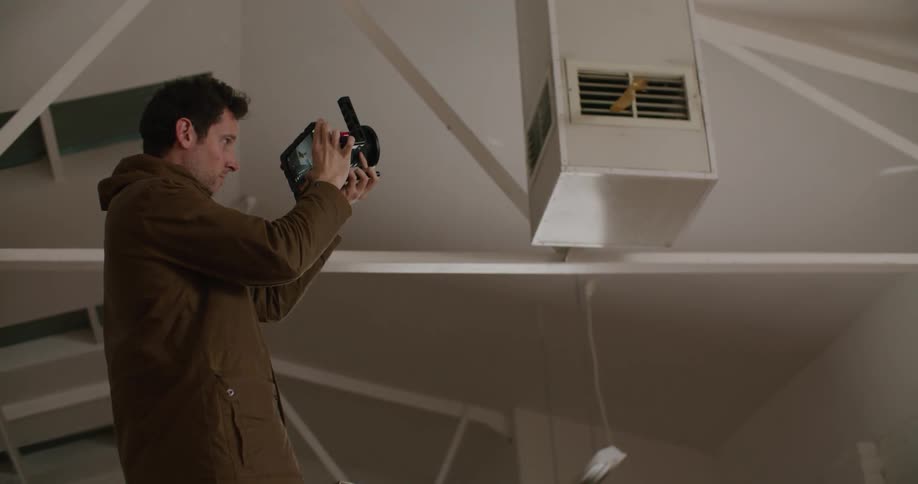 A man stands on a ladder and records a ribbon blowing out of an open air vent as another man watches from below