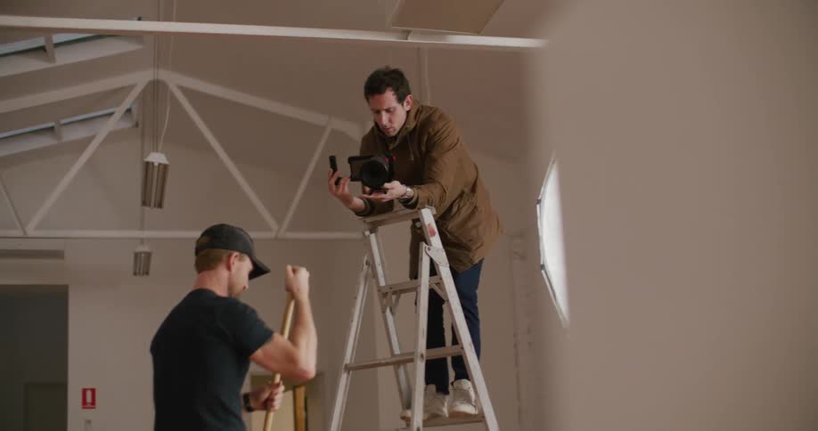 A photographer standing on a folding ladder prepares to take a photo of another man holding who is holding a broom