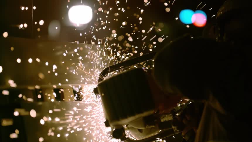 Sparks fly as person applies power tool to metal surface as colored lights form dots on glass