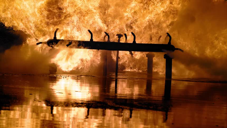 A sprinkler spraying water with fire and smoke in the background