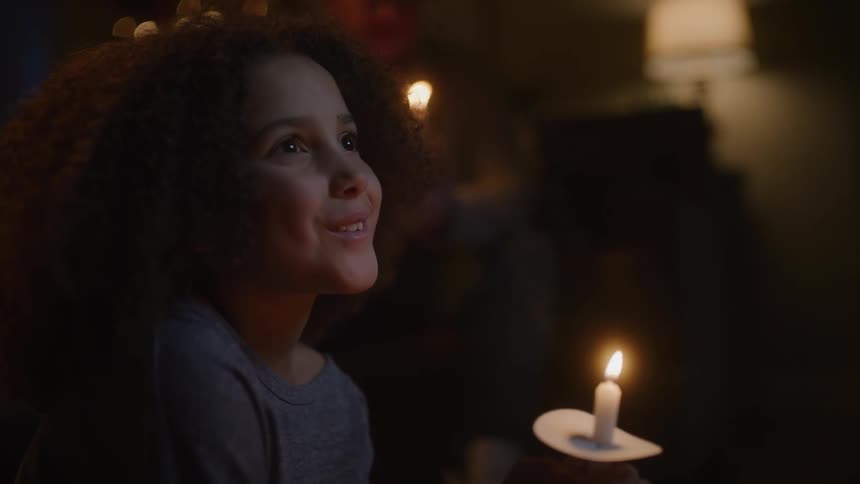 A girl in a room holding a candle with a man a lamp and a wall in the background