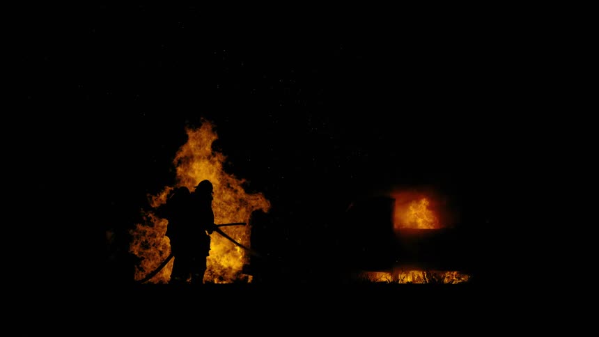 The silhouette of a firefighter can be seen in front of a raging fire at night